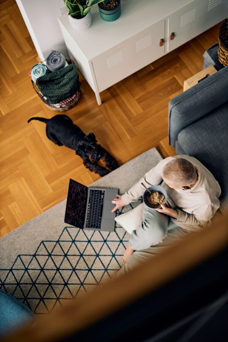 Top view of a senior woman sitting on the floor at home, eating breakfast, and using a laptop. Next to her is a dog trying to get attention.